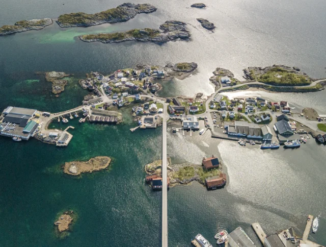 Aerial view of a coastal village with houses, docks, and small islands connected by a road. The sea surrounds the area, reflecting sunlight.