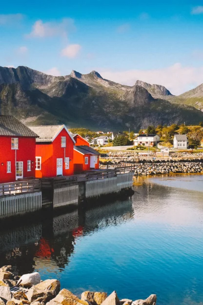A coastal scene with red wooden houses by a calm body of water, rocky shoreline, and mountains under a clear blue sky.