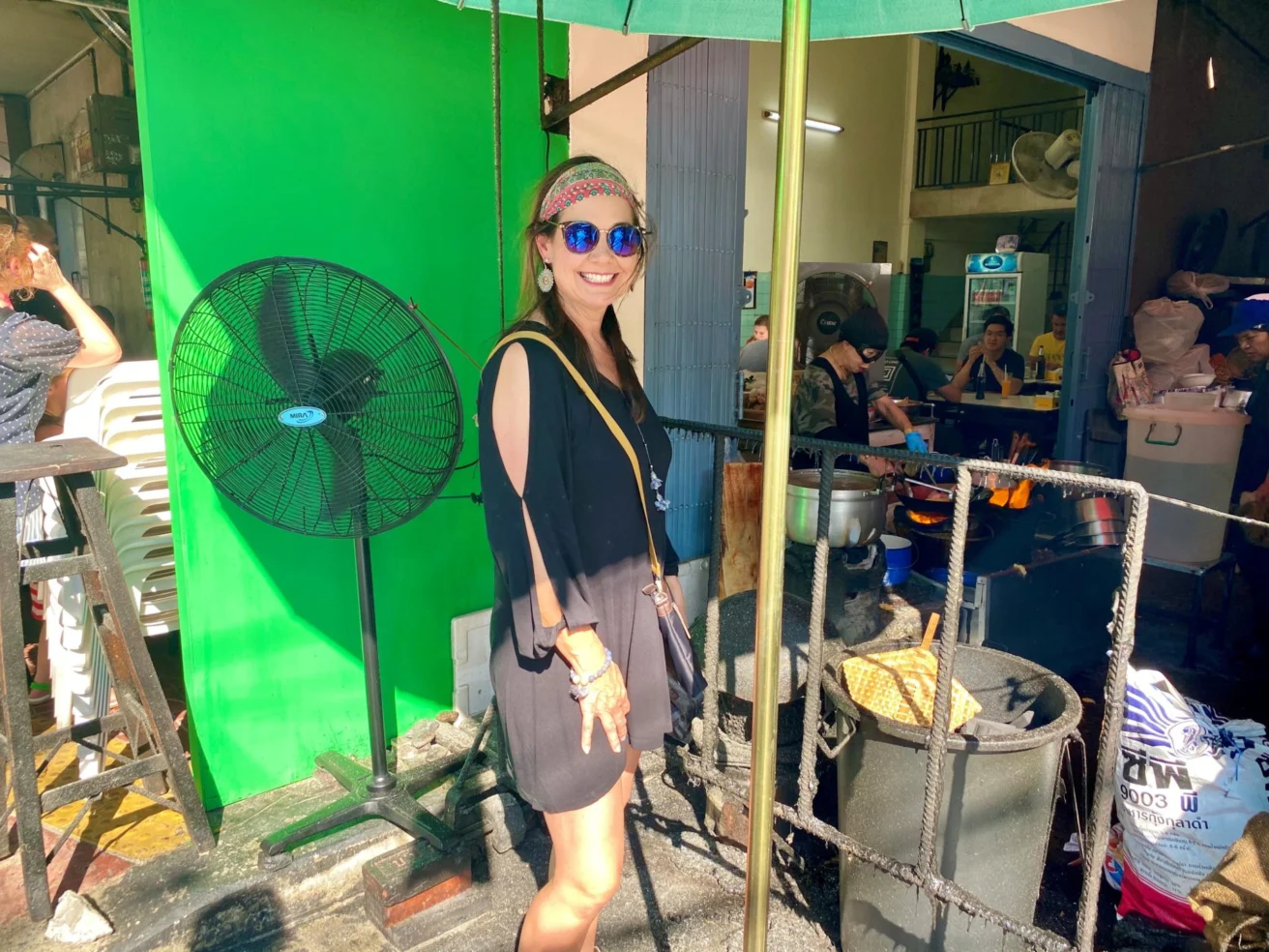 A person in sunglasses and a black dress stands smiling near a sidewalk food stall with a large fan nearby. Other people are eating in the background.