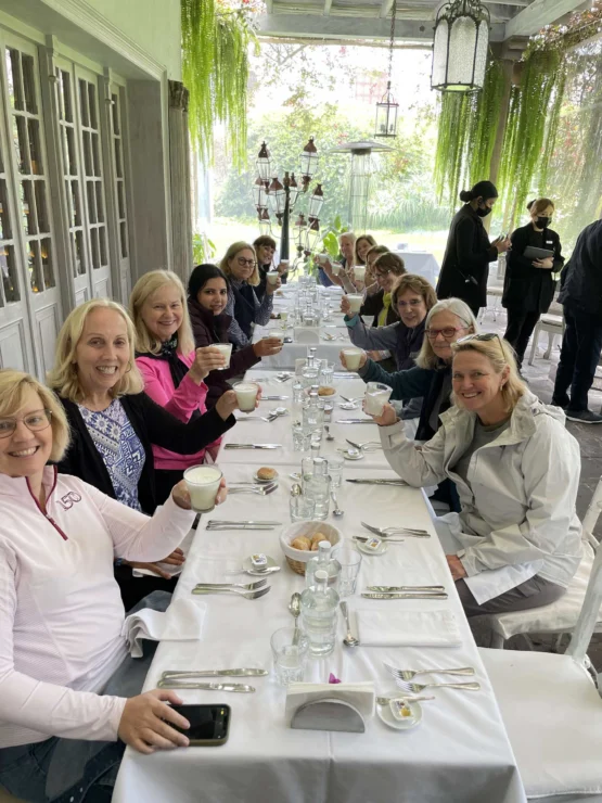 A group of people sitting at a long, white-covered table outdoors, holding drinks and smiling. The setting is surrounded by greenery and hanging plants.