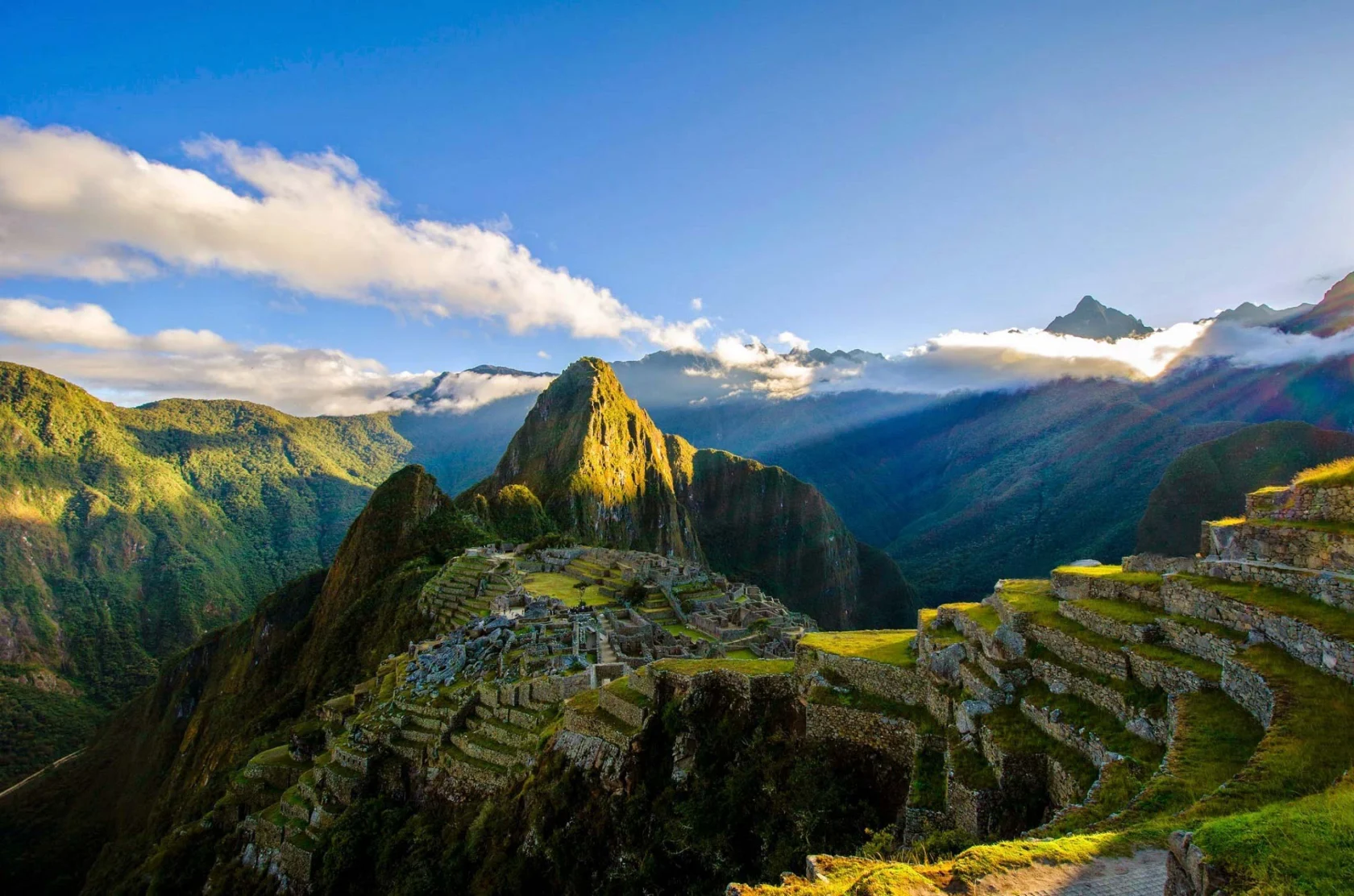 Ancient ruins of Machu Picchu on a mountain, surrounded by greenery and clouds under a clear sky.