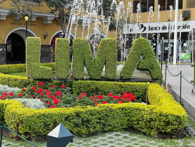 A decorative plant display spells "LIMA" in a garden with red flowers and small fountains, set against city buildings and a pigeon in the foreground.