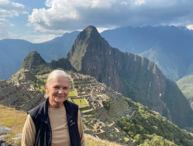 An individual stands in front of the ancient ruins of Machu Picchu with a mountain backdrop under a partly cloudy sky.