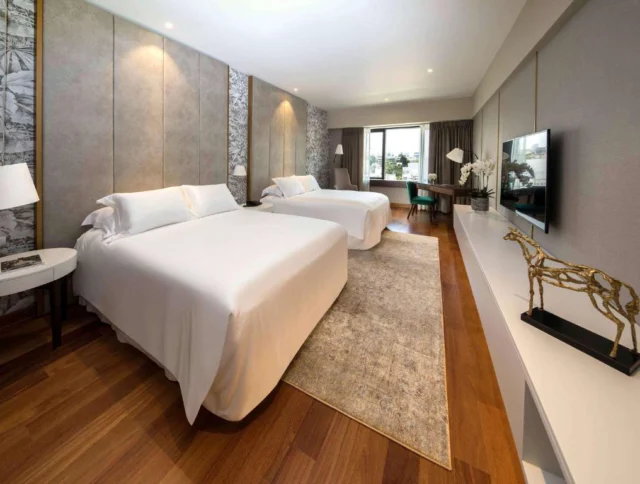 A modern hotel room with two double beds, wood flooring, a beige rug, a wall-mounted TV, a decorative giraffe sculpture, and a view through large windows.