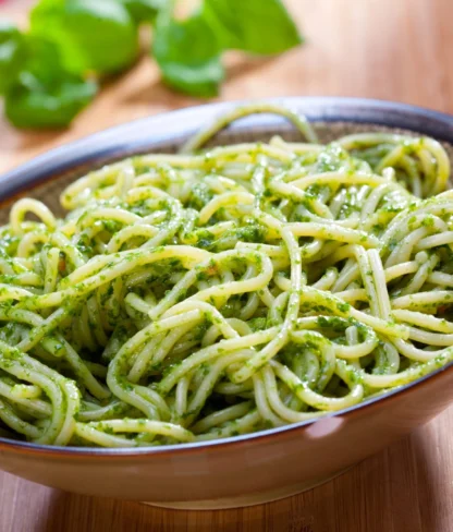 A bowl of spaghetti tossed with green pesto sauce, garnished with basil leaves, placed on a wooden surface.