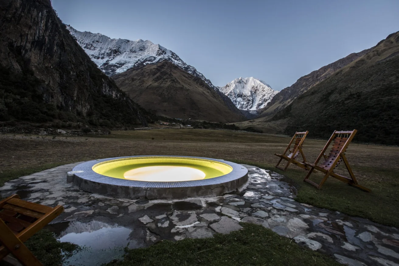 A round hot tub with glowing yellow light is set in a mountainous landscape with snow-capped peaks. Two wooden lounge chairs are positioned nearby.