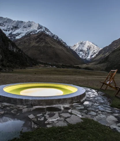 A round hot tub with glowing yellow light is set in a mountainous landscape with snow-capped peaks. Two wooden lounge chairs are positioned nearby.