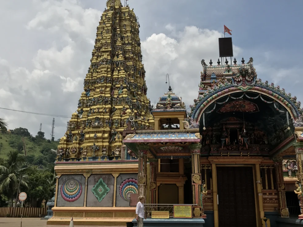A colorful Hindu temple with an ornate tower stands against a cloudy sky. A person is walking in the foreground.