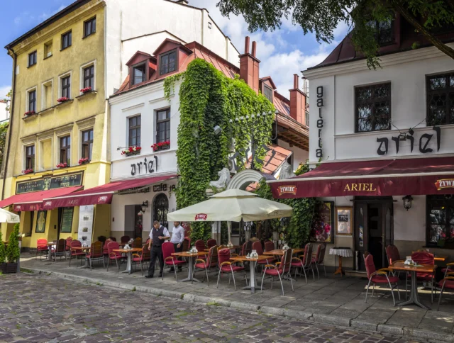 A street view of colorful, ivy-covered buildings with a cafe set up outside on a cobblestone path. Red chairs and umbrellas are arranged around tables, with a few people nearby.