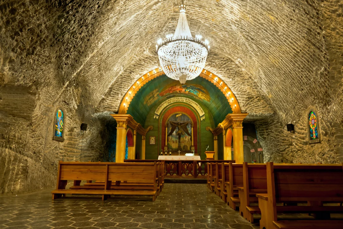 A chapel inside a salt mine, featuring wooden pews, an altar, a colorful mural, stained glass windows, and a large chandelier hanging from the arched ceiling.
