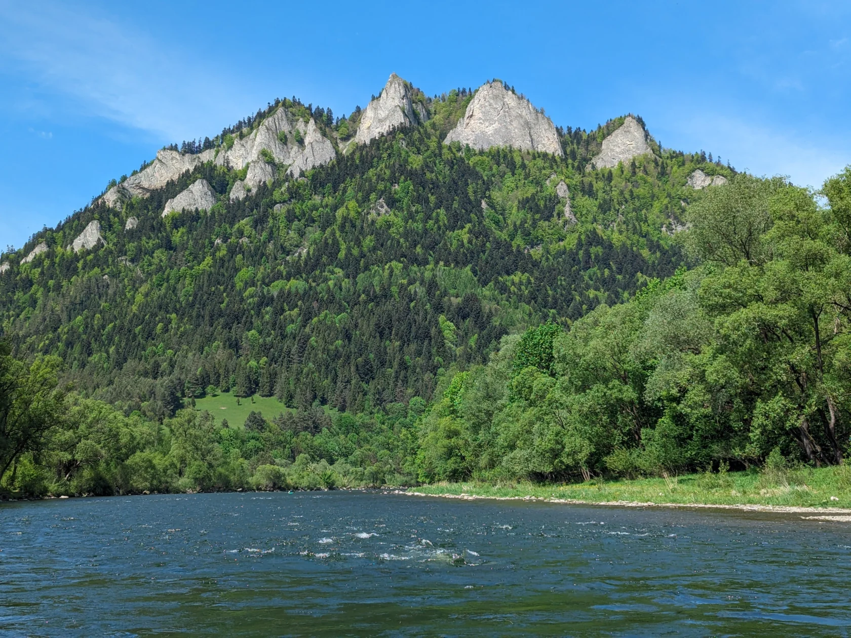A clear river flows in the foreground with a green, tree-covered mountain and sharp peaks under a blue sky in the background.