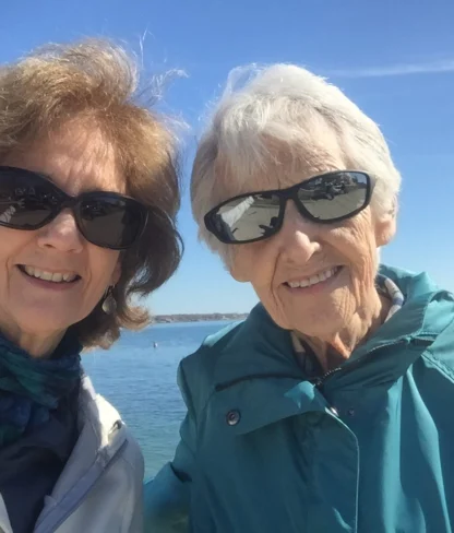 Two women wearing sunglasses pose for a selfie by the water on a sunny day.