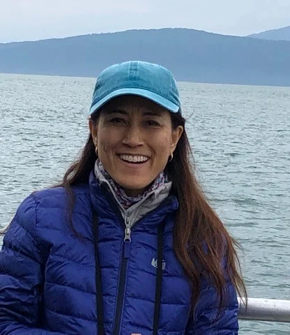 A person wearing a blue jacket and cap stands smiling in front of a body of water with mountains in the background.