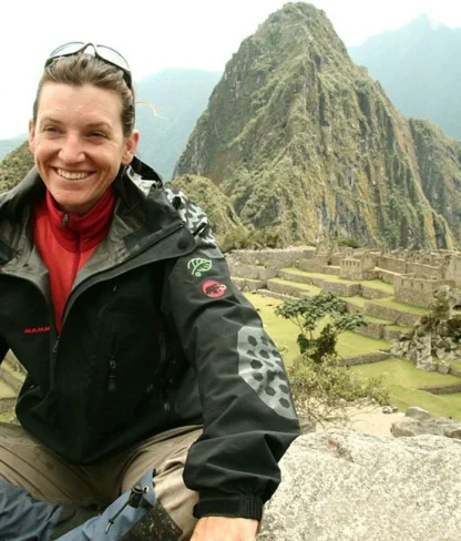 A person sits on a rock at Machu Picchu, smiling, with mountains and ancient ruins in the background.