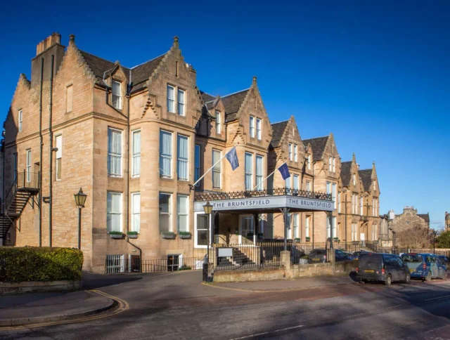 Exterior of a stone building with multiple windows, steep roofs, and a sign reading "The Bruntsfield." Cars are parked along the street under a clear blue sky.