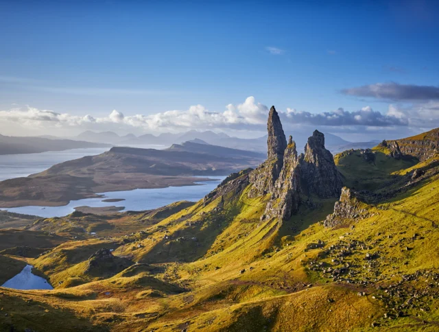 Mountain landscape with rock formations and rolling hills under a clear blue sky, overlooking a body of water in the distance.