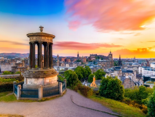 View of Edinburgh from Calton Hill at sunset, featuring the Dugald Stewart Monument in the foreground and the cityscape with colorful skies in the background.