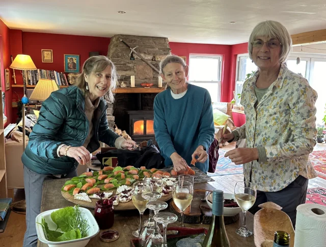 Three women prepare food at a table in a cozy living room with a fireplace. There are appetizers, wine glasses, and a bottle of wine on the table.