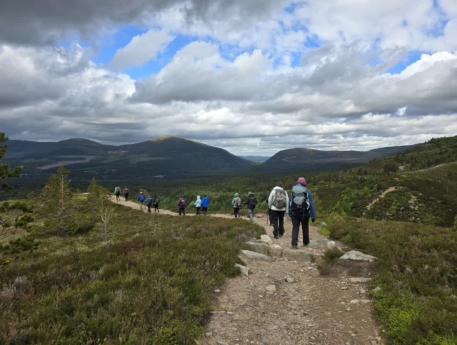 A group of hikers with backpacks walks along a mountainous trail under a cloudy sky.