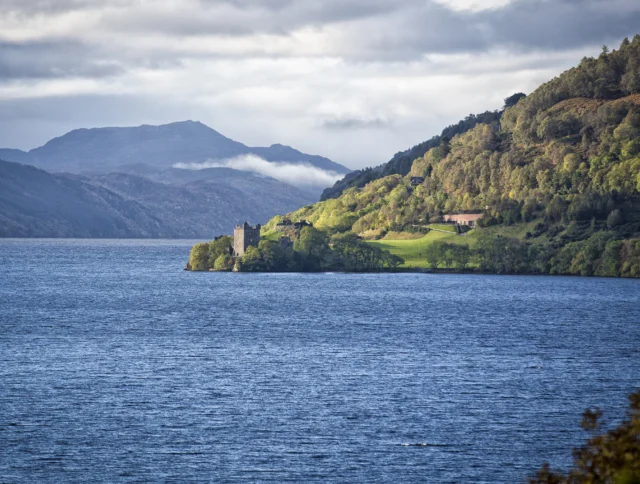 A calm lake with a historic castle on a grassy hill, surrounded by forested mountains under a partly cloudy sky.