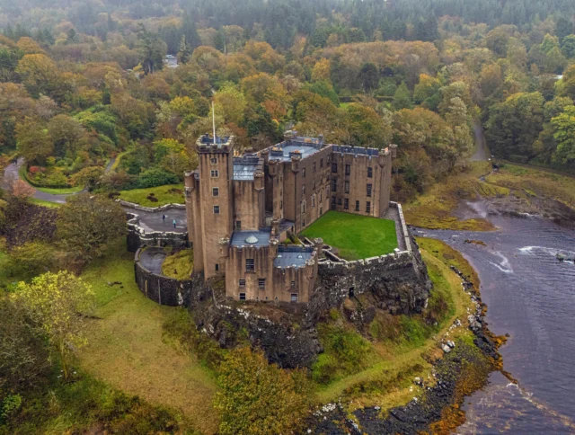 Aerial view of a historic castle surrounded by trees and a river, with a winding road leading to its entrance.