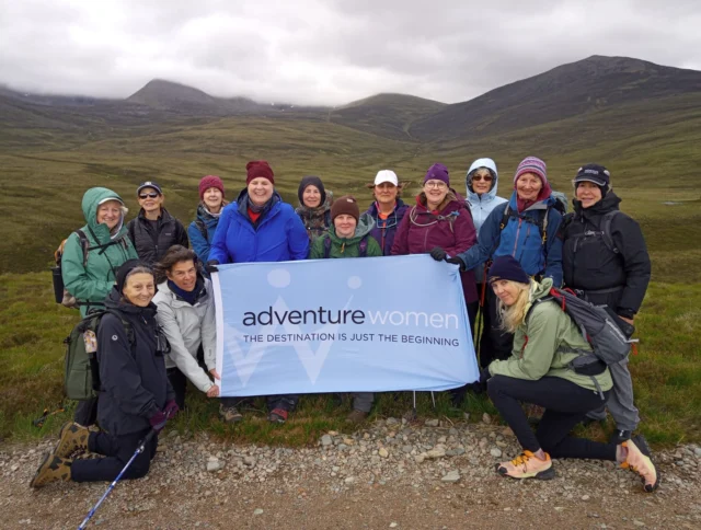 A group of women outdoors holding a banner that reads "adventure women, the destination is just the beginning," with hills in the background.
