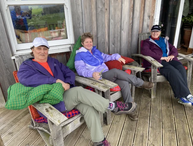 Three people sit on wooden chairs on a deck, wearing outdoor jackets and caps, with a cabin wall behind them.