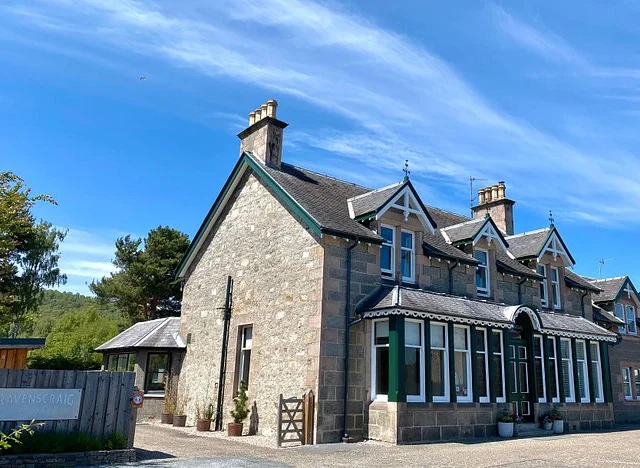 Stone house with steep roofs, multiple chimneys, and large windows under a blue sky. A sign near the entrance reads "Ravenscraig." Trees and clouds in the background.