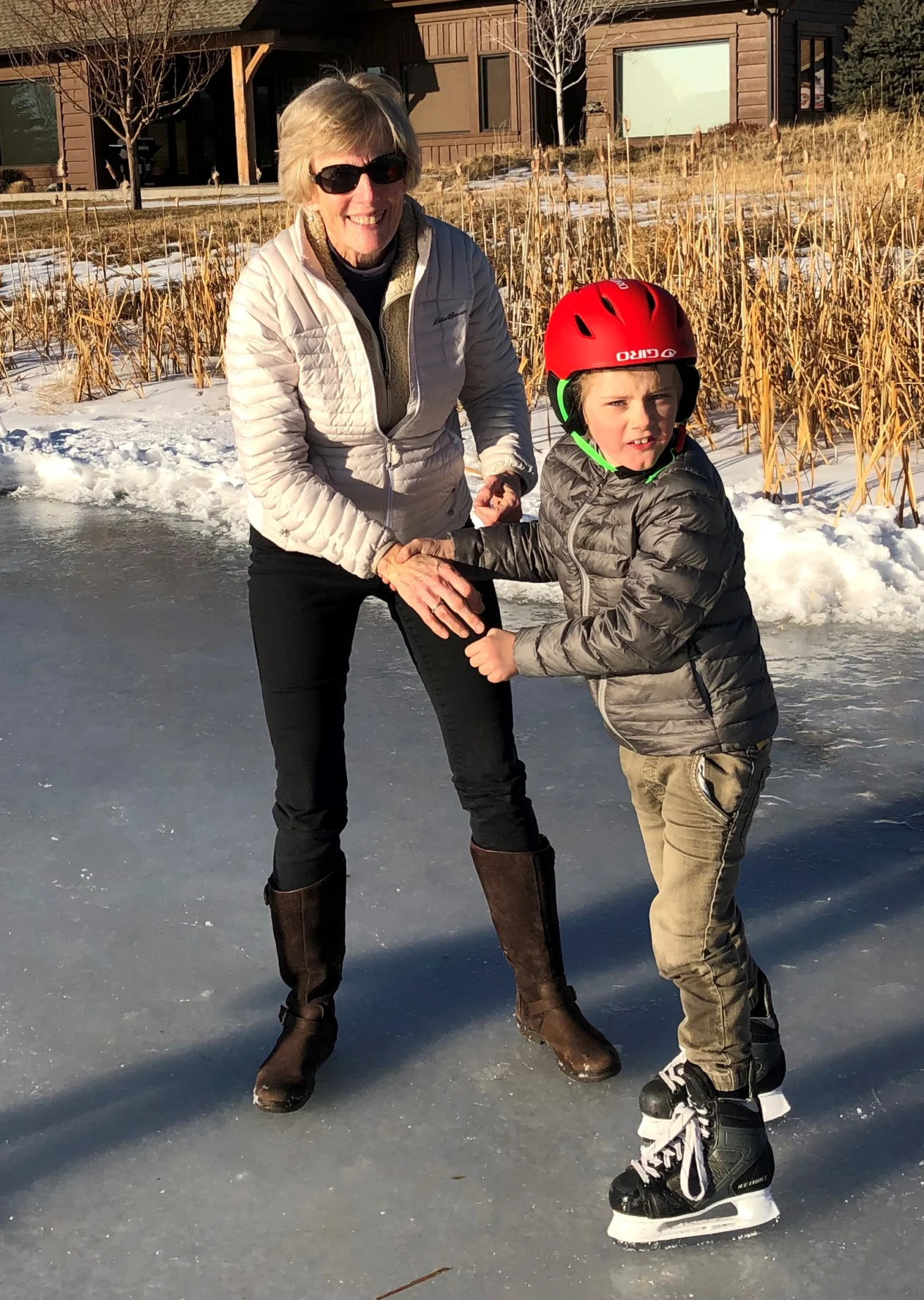 A woman and a child in a red helmet ice skating together on a frozen outdoor pond in a sunny winter setting.