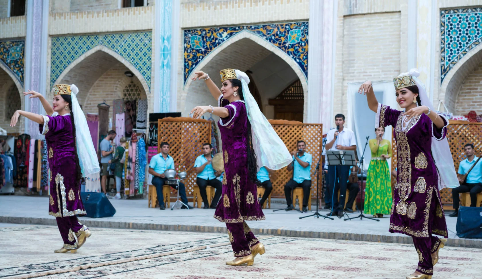 Three women in traditional purple attire perform a cultural dance in an ornate courtyard, with musicians playing in the background.
