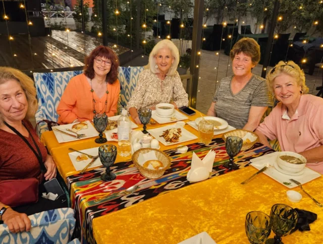 Five women sitting at a restaurant table with partially eaten meals, smiling at the camera. The background is decorated with string lights.