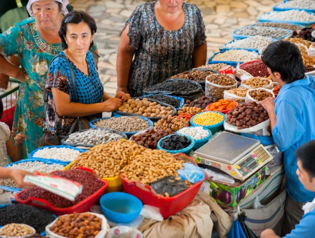 People selling and buying dried fruits, nuts, and beans at a market stall. There are large baskets filled with various colorful items.