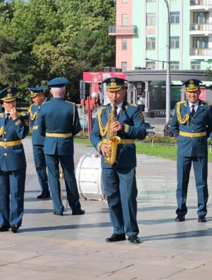 A marching band in military uniforms performs outdoors with various instruments, including tubas, saxophones, and a drum, on a paved area with buildings and trees in the background.