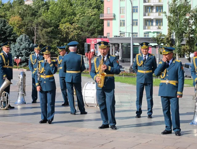 A marching band in military uniforms performs outdoors with various instruments, including tubas, saxophones, and a drum, on a paved area with buildings and trees in the background.