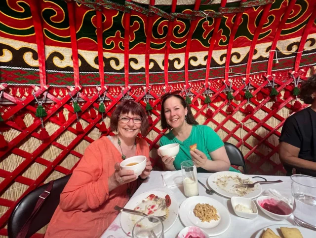Two women are sitting at a table inside a colorful yurt, smiling and holding bowls. The table has finished dishes and drinks.