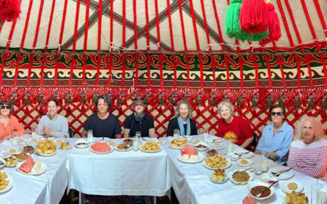 A group of people seated around a long, food-filled table inside a yurt with colorful patterned walls and ceiling.