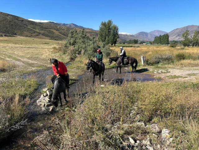 Three people on horseback cross a rocky stream in a grassy, mountainous landscape under a clear blue sky.