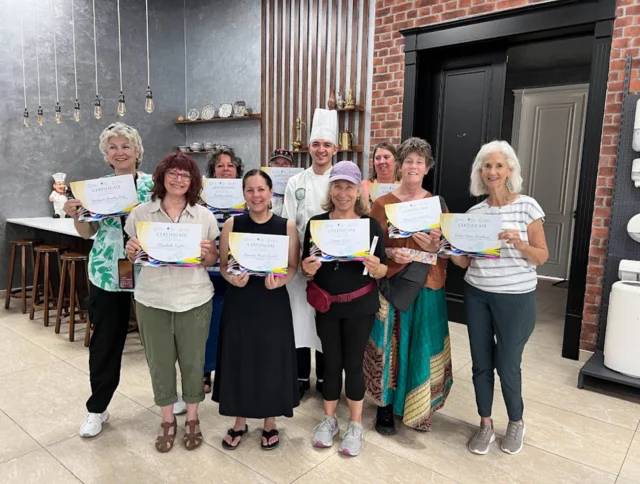 A group of eight smiling people holding certificates pose with a chef in a kitchen setting.