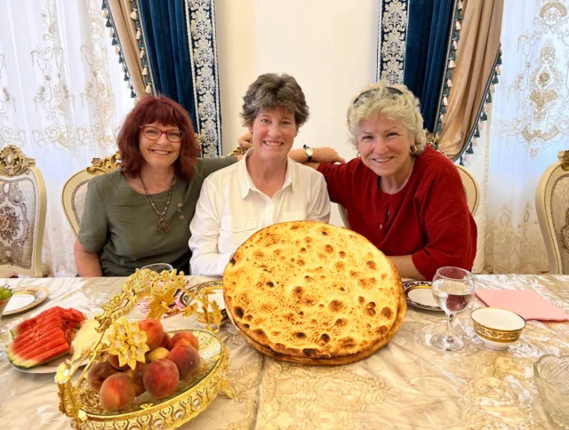 Three women smiling at a dining table with a large round bread, a plate of peaches, and a bowl of watermelon slices. Elegant chairs and curtains in the background.