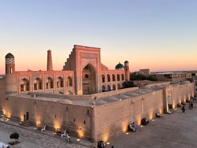 Sunset view of an ancient brick building with turquoise domes and intricate archways, surrounded by a courtyard and distant cityscape.