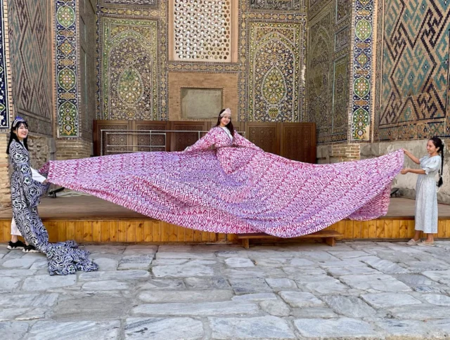 Three people display an exceptionally long, colorful dress with intricate patterns, standing against a backdrop of ornate architectural details.