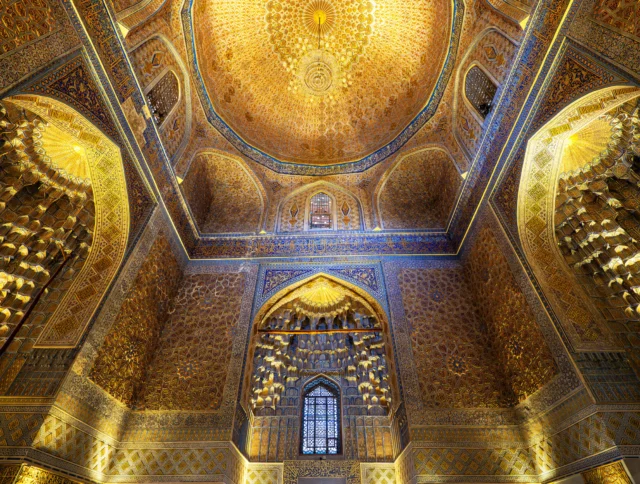 Ornate interior of an intricately decorated mosque dome, featuring gold and blue patterns with a central chandelier and arched windows.