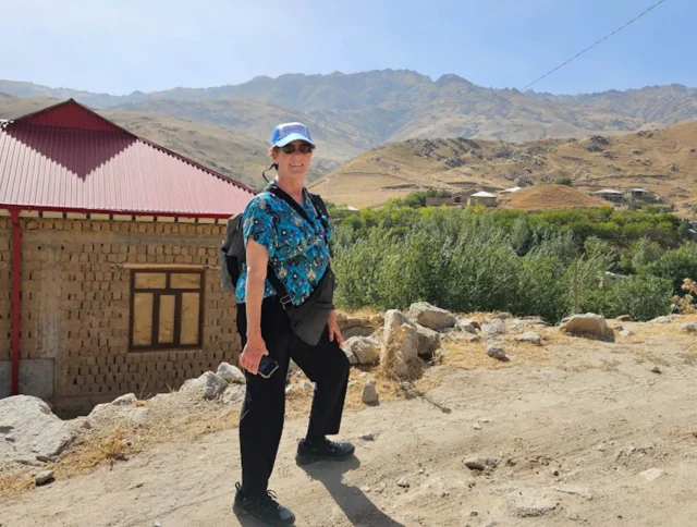 A person wearing a cap and sunglasses stands on a dirt path near a stone house. Hilly terrain and scattered buildings are visible in the background.