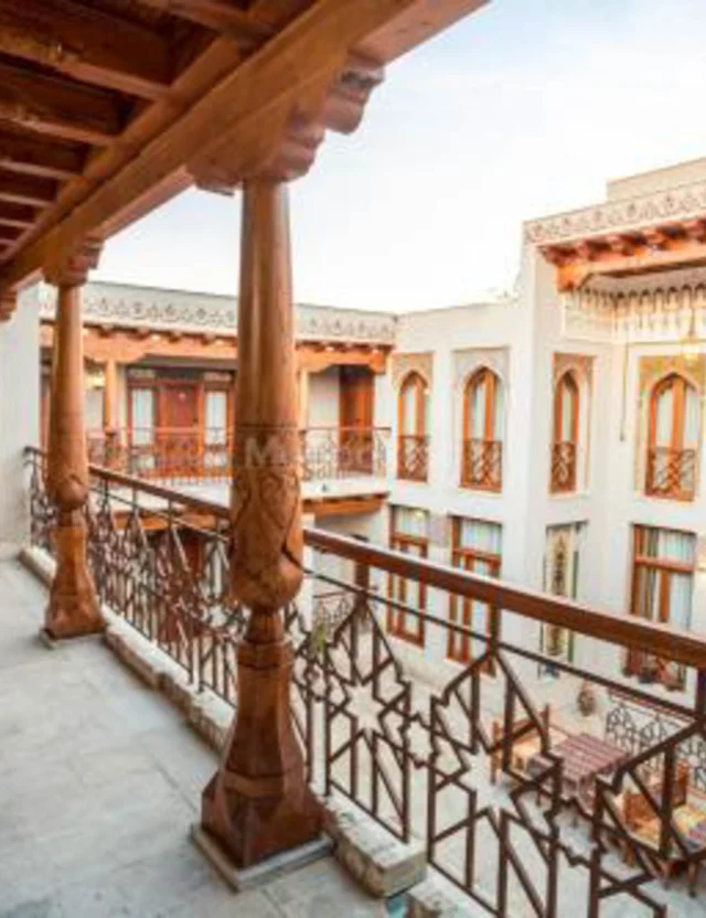 A hotel corridor with wooden railings and columns overlooks a central courtyard surrounded by two-story buildings featuring numerous windows with decorative wooden details.