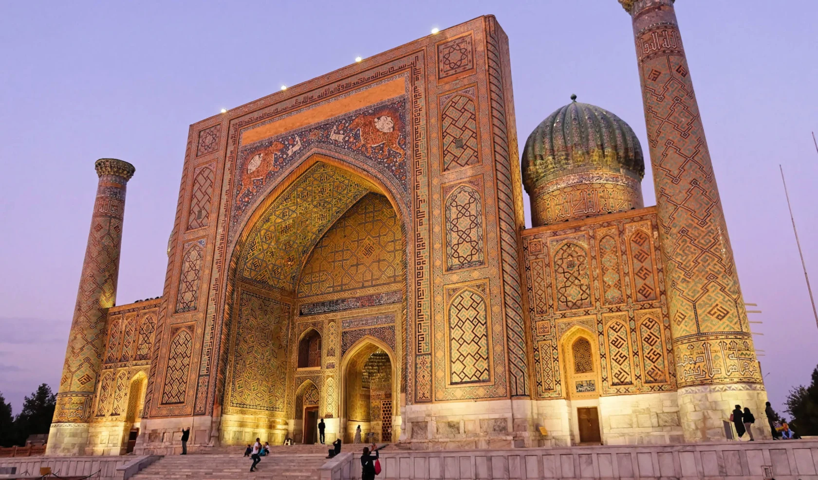A large, ornately decorated building with a domed roof and intricate patterns, featuring arches and a minaret, against a twilight sky.