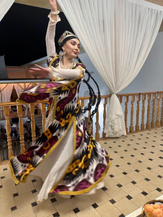 A woman in traditional attire spins gracefully, her colorful dress flowing, on a checkered floor with draped curtains in the background.