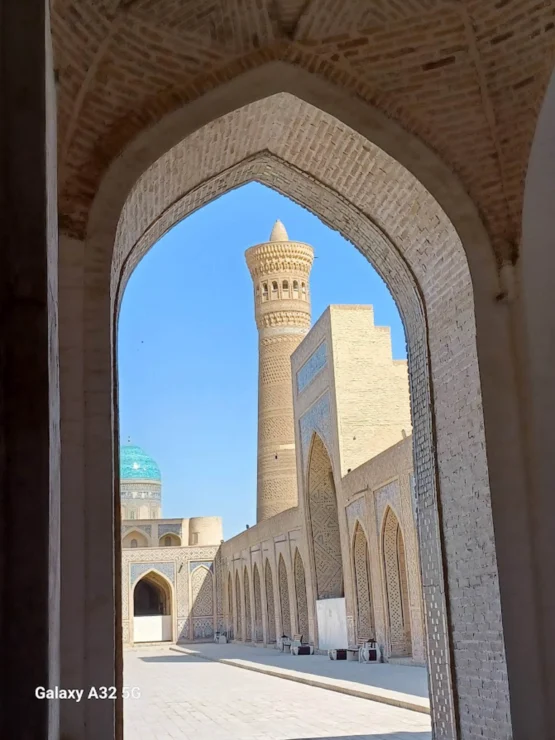View through an archway of a historic courtyard with a tall minaret and domed buildings under a clear blue sky.