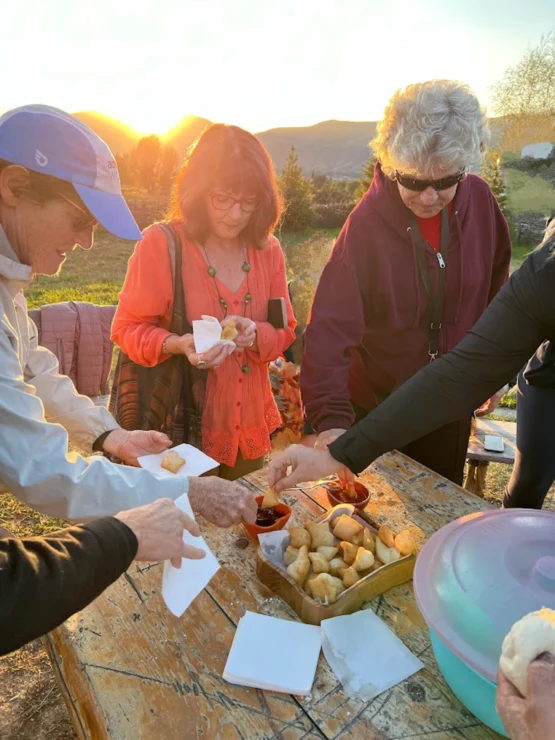 Group of people outdoors enjoying snacks from a table with various food items. Sunlight is visible in the background.