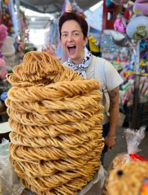 Person excitedly poses behind a large stack of twisted, golden-brown fried snacks at an outdoor market. Balloons and colorful items are displayed in the background.