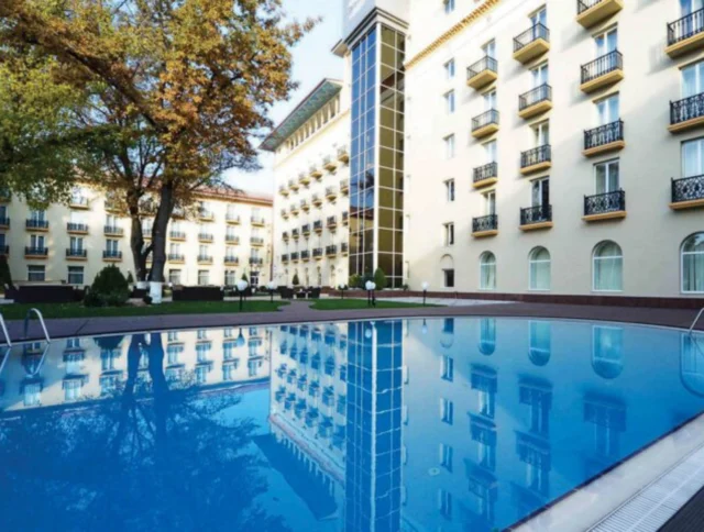 Outdoor pool with a clear blue surface reflecting a multi-story building with balconies and large windows, surrounded by trees and a neatly paved area.